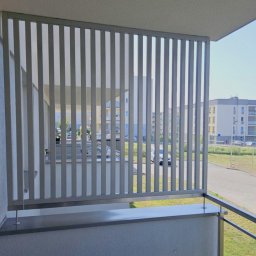 EXPO Kawałkowski Krzysztof - Nowoczesna balustrada balkonowa z pionowych, szarych paneli, zapewniająca prywatność i przepuszczająca światło. Widok na osiedle z balkonu.