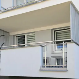 Nowoczesny balkon z szklaną balustradą i metalowymi panelami, wyposażony w stolik i krzesła. Widok z zewnątrz na jasną elewację budynku.
