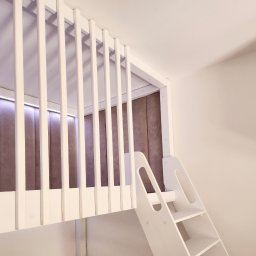 EXPO Kawałkowski Krzysztof - Białe, drewniane łóżko piętrowe z drabinką, zintegrowane z tapicerowanym panelem ściennym w pokoju dziecięcym. Nowoczesny design, minimalistyczny styl.