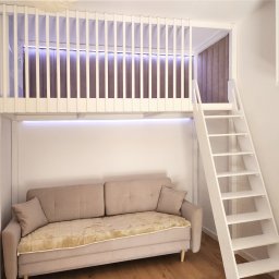 EXPO Kawałkowski Krzysztof - Biały pokój dziecięcy z antresolą i drabinką. Pod antresolą beżowa sofa z poduszkami. Górna część antresoli zabezpieczona białymi szczebelkami.