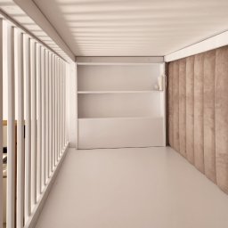 EXPO Kawałkowski Krzysztof - Minimalistyczna garderoba z białymi półkami i beżowym panelem ściennym, widok z perspektywy wejścia przez białą, ażurową balustradę.