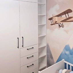 EXPO Kawałkowski Krzysztof - Biała szafa z czarnymi uchwytami w pokoju dziecięcym z motywem samolotu na ścianie i białym łóżeczkiem. Widok na półki szafy i szuflady.