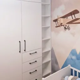 Biała szafa z czarnymi uchwytami w pokoju dziecięcym z motywem samolotu na ścianie i białym ł&oacute;żeczkiem. Widok na p&oacute;łki szafy i szuflady.