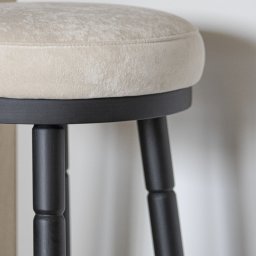 EXPO Kawałkowski Krzysztof - Detale tapicerowanego stołka barowego z ciemnymi nogami, na tle jasnej ściany. Widoczny fragment siedziska i nogi stołka.