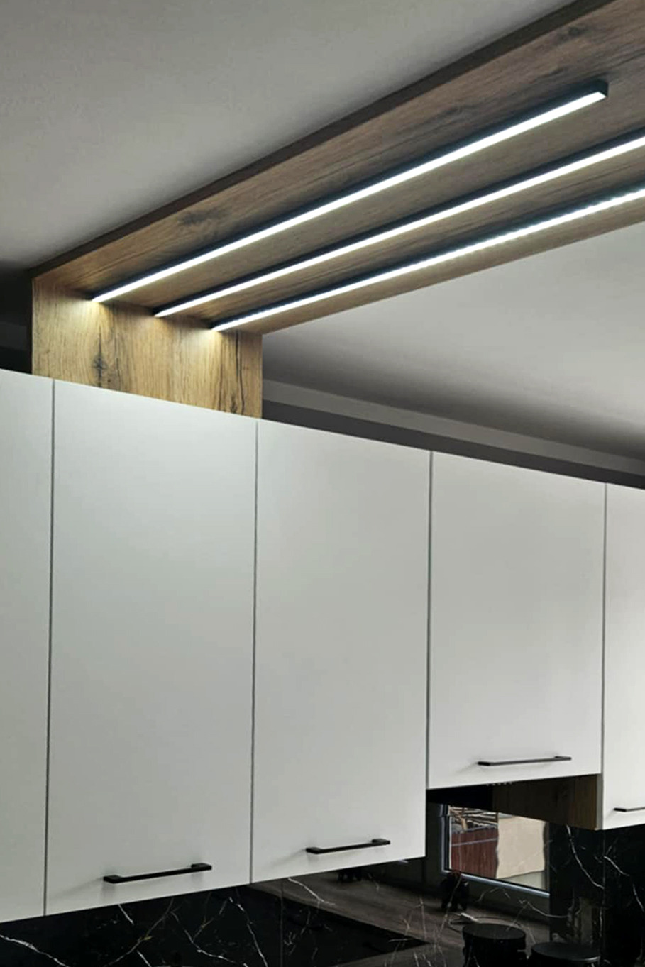 Nowoczesna kuchnia z białymi szafkami i blatem imitującym czarny marmur. Oświetlenie LED w drewnianej zabudowie nad szafkami dodaje wnętrzu elegancji.