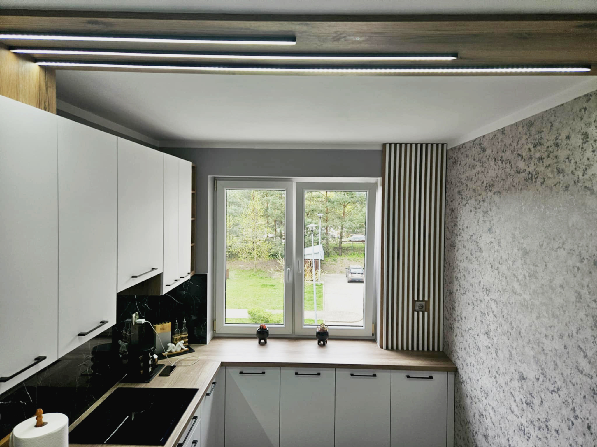 Nowoczesna kuchnia z białymi szafkami, czarnym blatem imitującym marmur i oknem z widokiem na zieleń. Oświetlenie LED podkreśla drewniane elementy wykończenia.