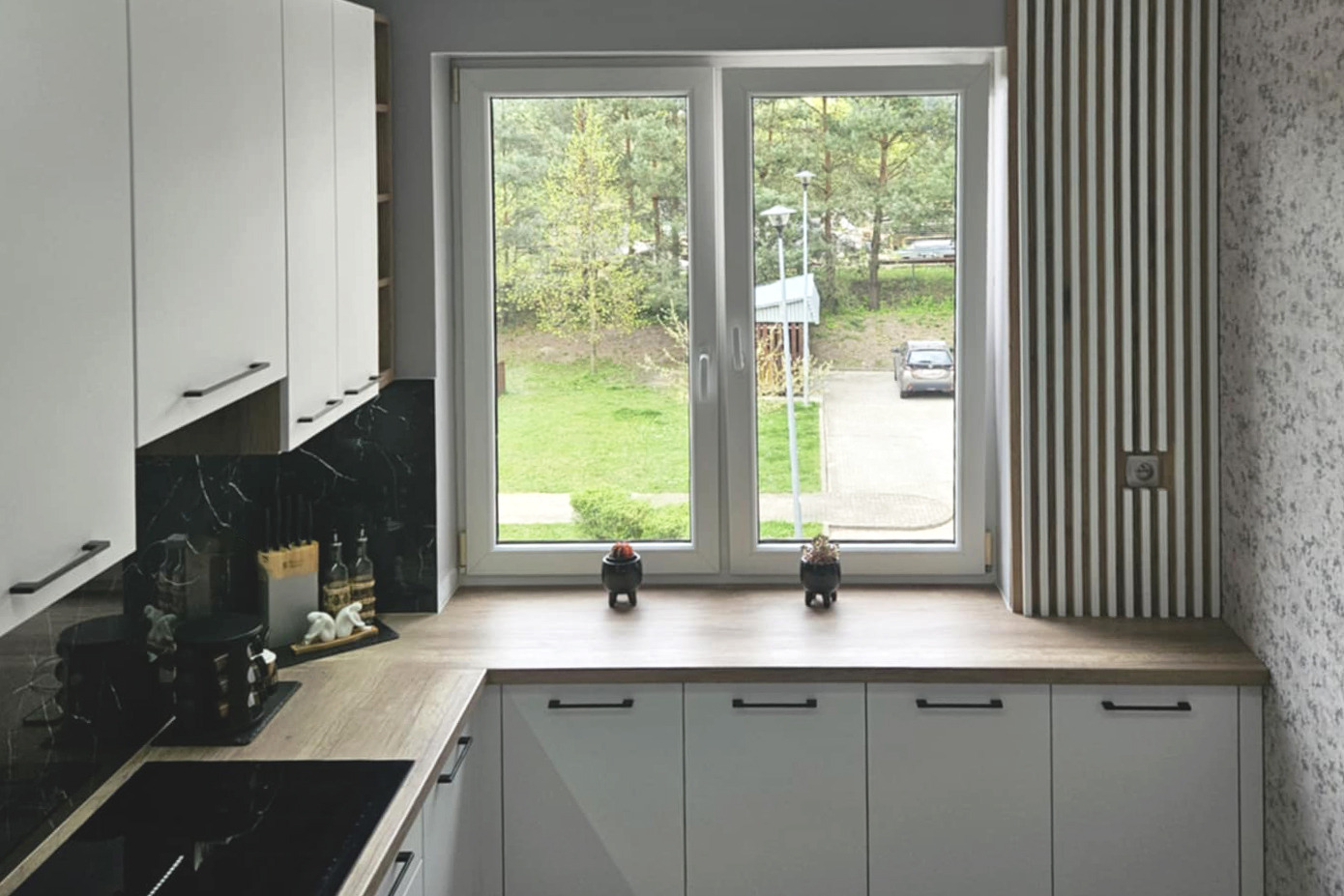 Jasna kuchnia z oknem na ogród. Białe szafki z czarnymi uchwytami, drewniany blat i czarny panel. Dekoracje okienne i widok na zieleń.