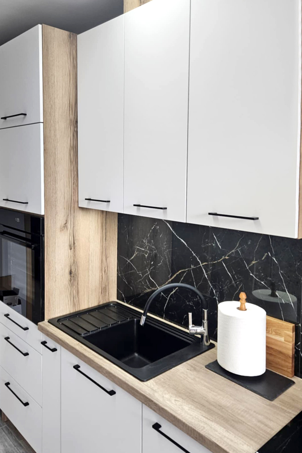 Nowoczesna kuchnia z białymi szafkami, drewnianymi akcentami i czarnym blatem z marmurkowym wzorem. Czarne, podłużne uchwyty i czarny zlew.