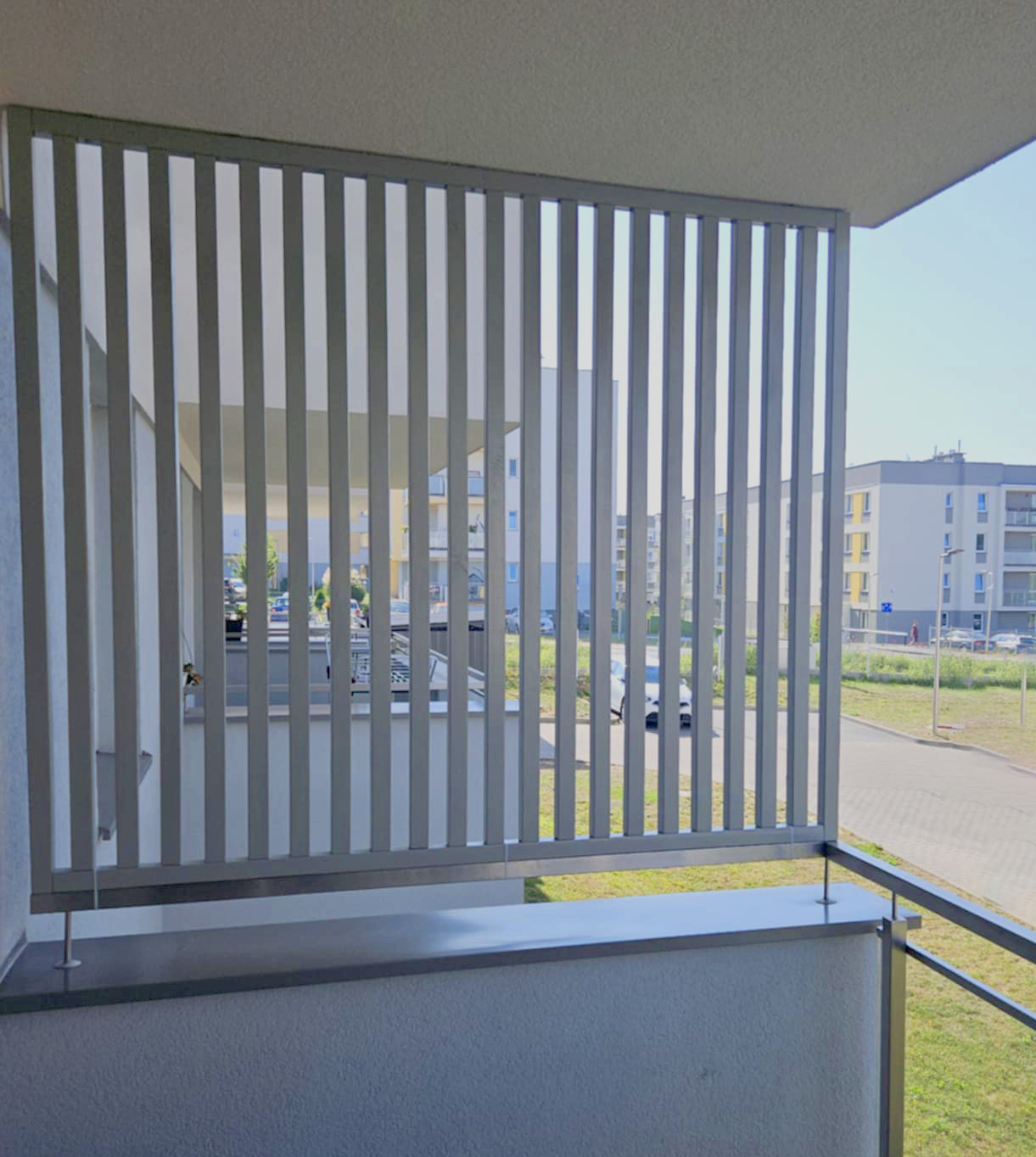 Nowoczesna balustrada balkonowa z pionowych, szarych paneli, zapewniająca prywatność i przepuszczająca światło. Widok na osiedle z balkonu.