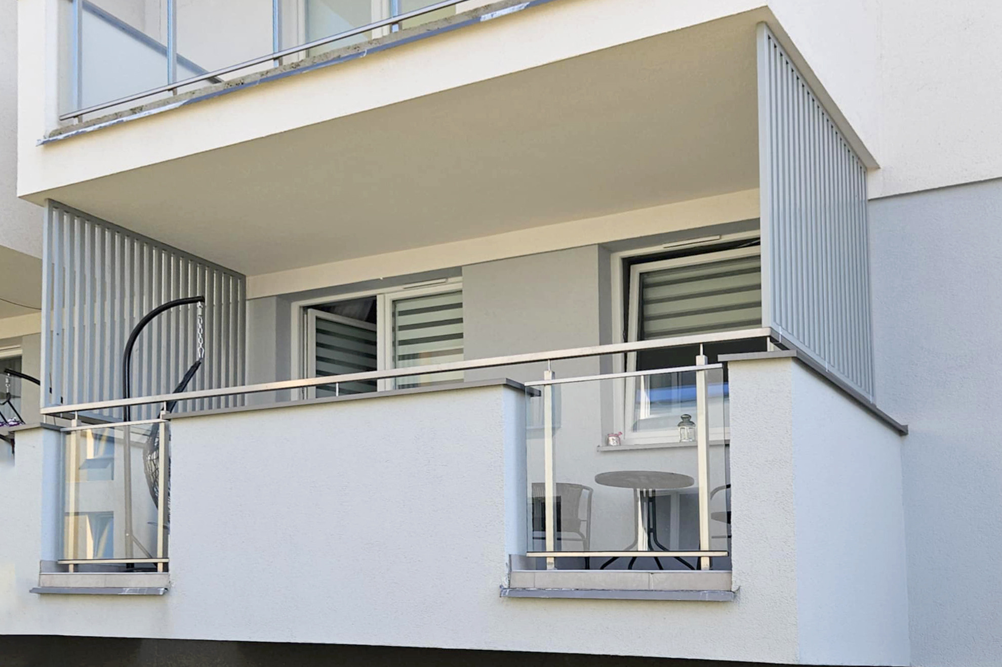 Nowoczesny balkon z szklaną balustradą i metalowymi panelami, wyposażony w stolik i krzesła. Widok z zewnątrz na jasną elewację budynku.