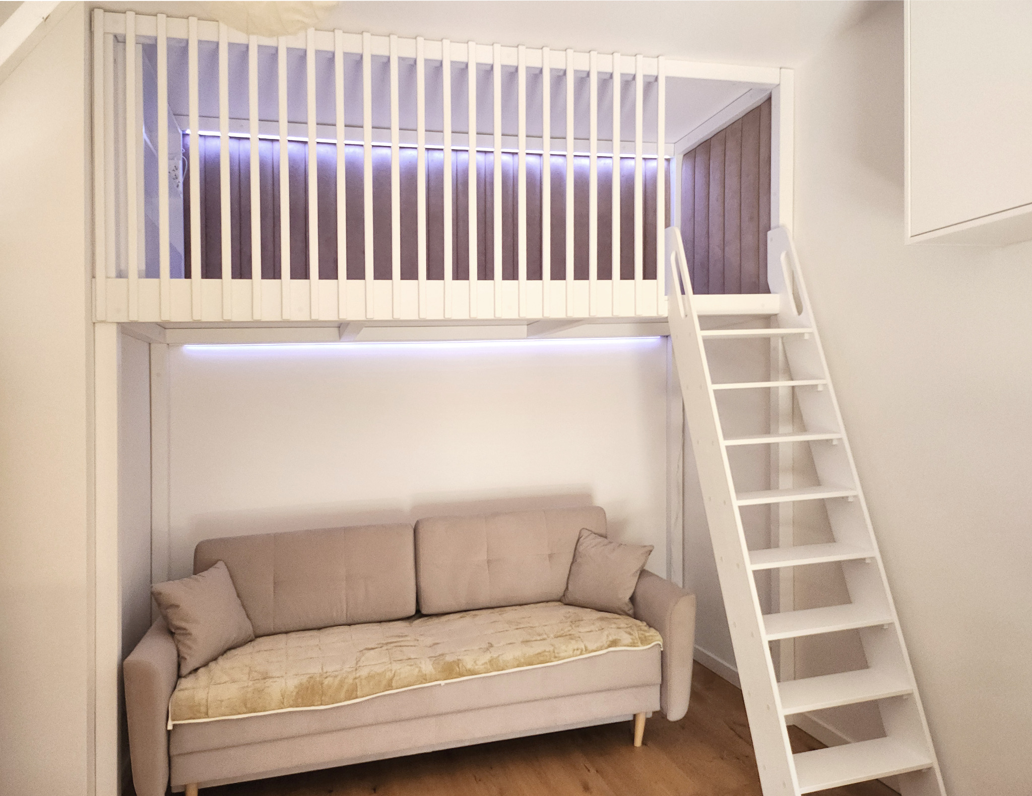 Biały pokój dziecięcy z antresolą i drabinką. Pod antresolą beżowa sofa z poduszkami. Górna część antresoli zabezpieczona białymi szczebelkami.