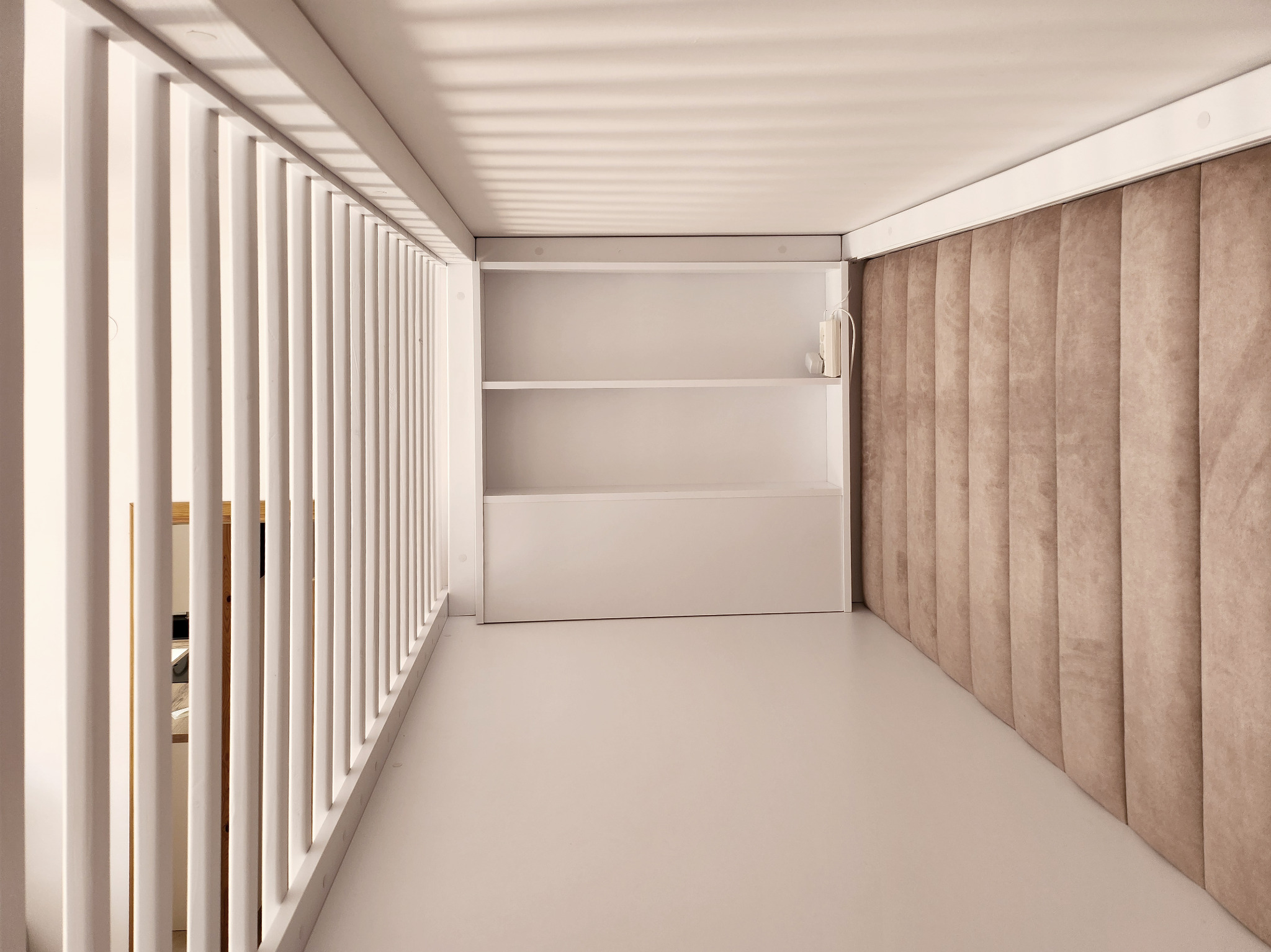 Minimalistyczna garderoba z białymi półkami i beżowym panelem ściennym, widok z perspektywy wejścia przez białą, ażurową balustradę.