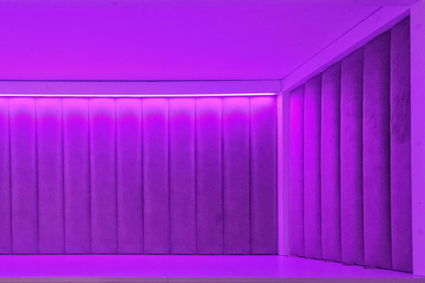 Nowoczesna ściana z paneli tapicerowanych w odcieniach fioletu, oświetlona listwą LED. Minimalistyczny design z pionowymi przeszyciami.