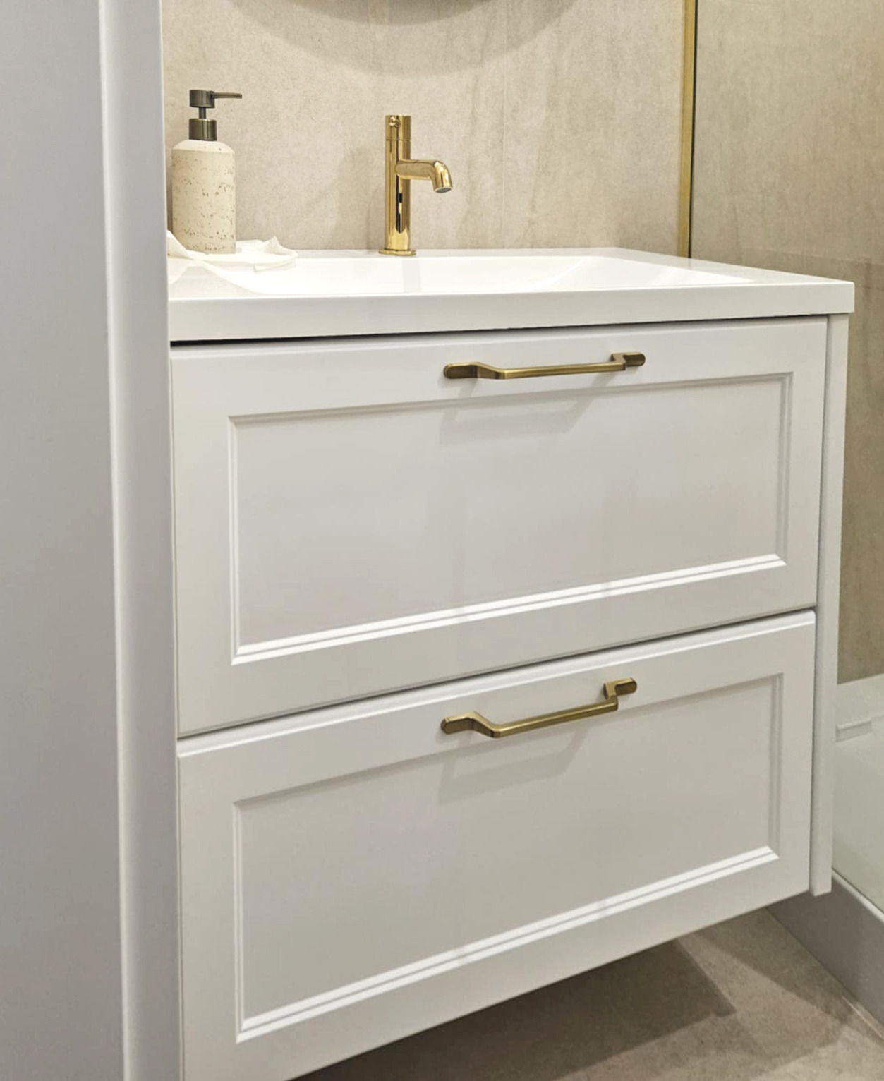 Biała szafka łazienkowa z dwiema szufladami i złotymi uchwytami, zwieńczona umywalką z baterią w kolorze złota. Na blacie dozownik mydła w odcieniach beżu.