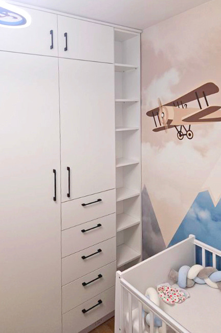 Biała szafa z czarnymi uchwytami w pokoju dziecięcym z motywem samolotu na ścianie i białym łóżeczkiem. Widok na półki szafy i szuflady.