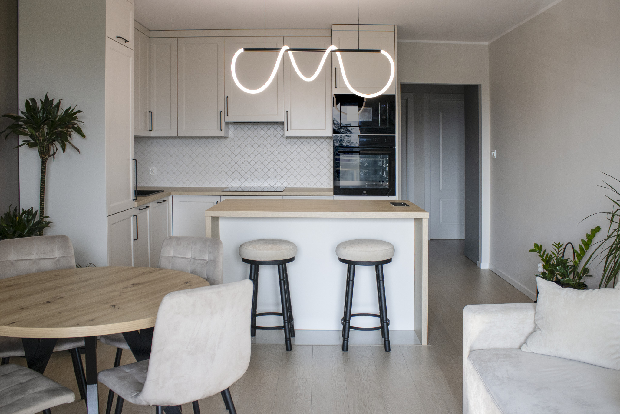 Jasna kuchnia z wyspą i nowoczesnym oświetleniem. Białe szafki, drewniany blat, okrągły stół z krzesłami i sofa. Całość w neutralnej kolorystyce.