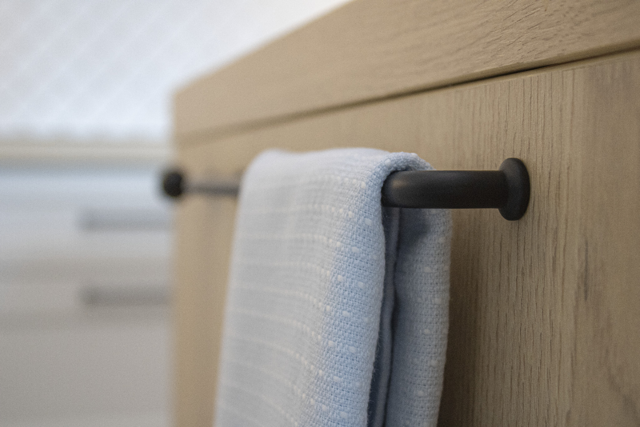 Fragment szafki z drewnianym frontem i czarnym relingiem, na którym wisi jasnoniebieski ręcznik. Ujęcie z bliska, z rozmytym tłem.
