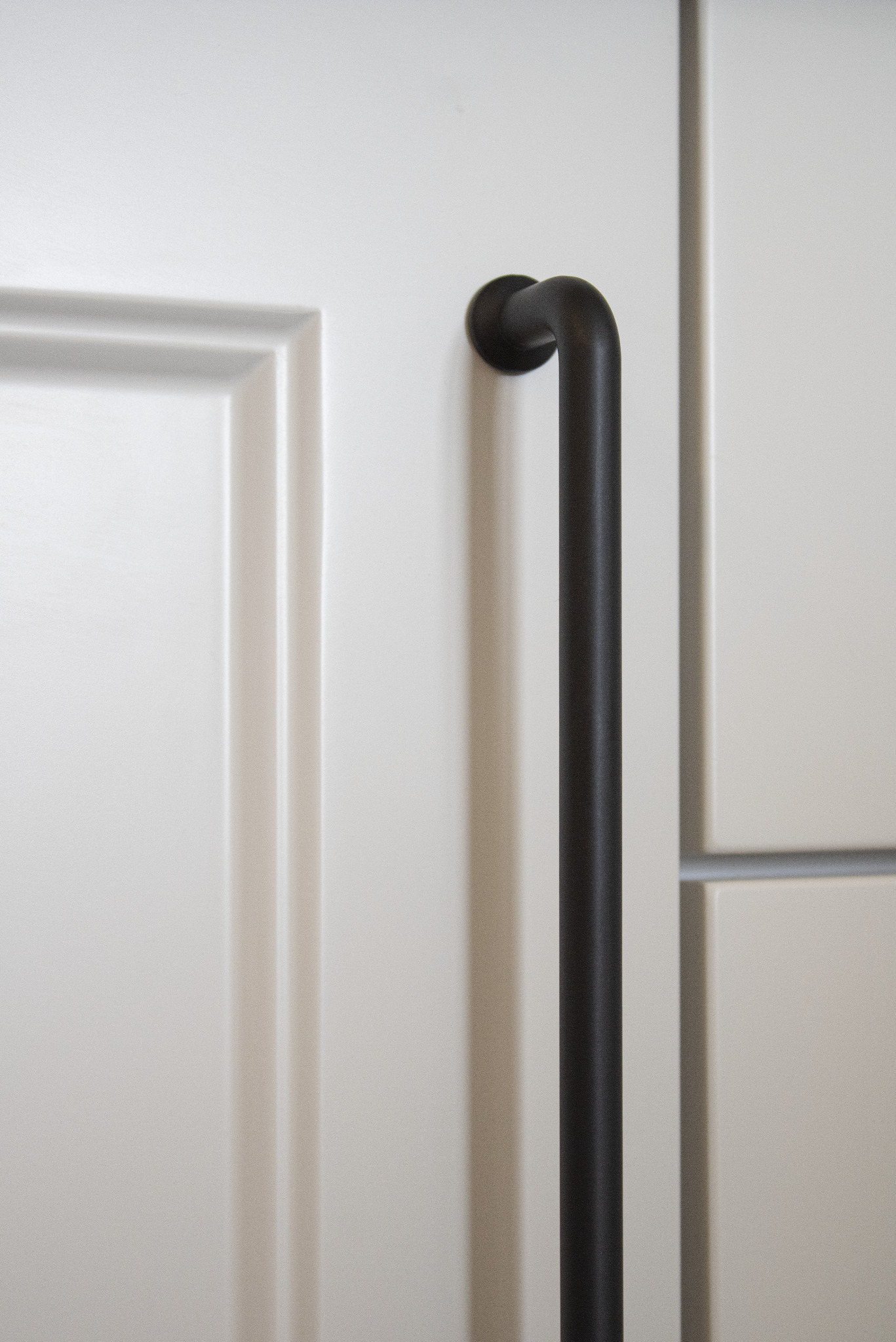 Biały front szafki z frezowaniem i czarnym, pionowym uchwytem rurowym. Minimalistyczny design, gładkie powierzchnie i subtelne światło.