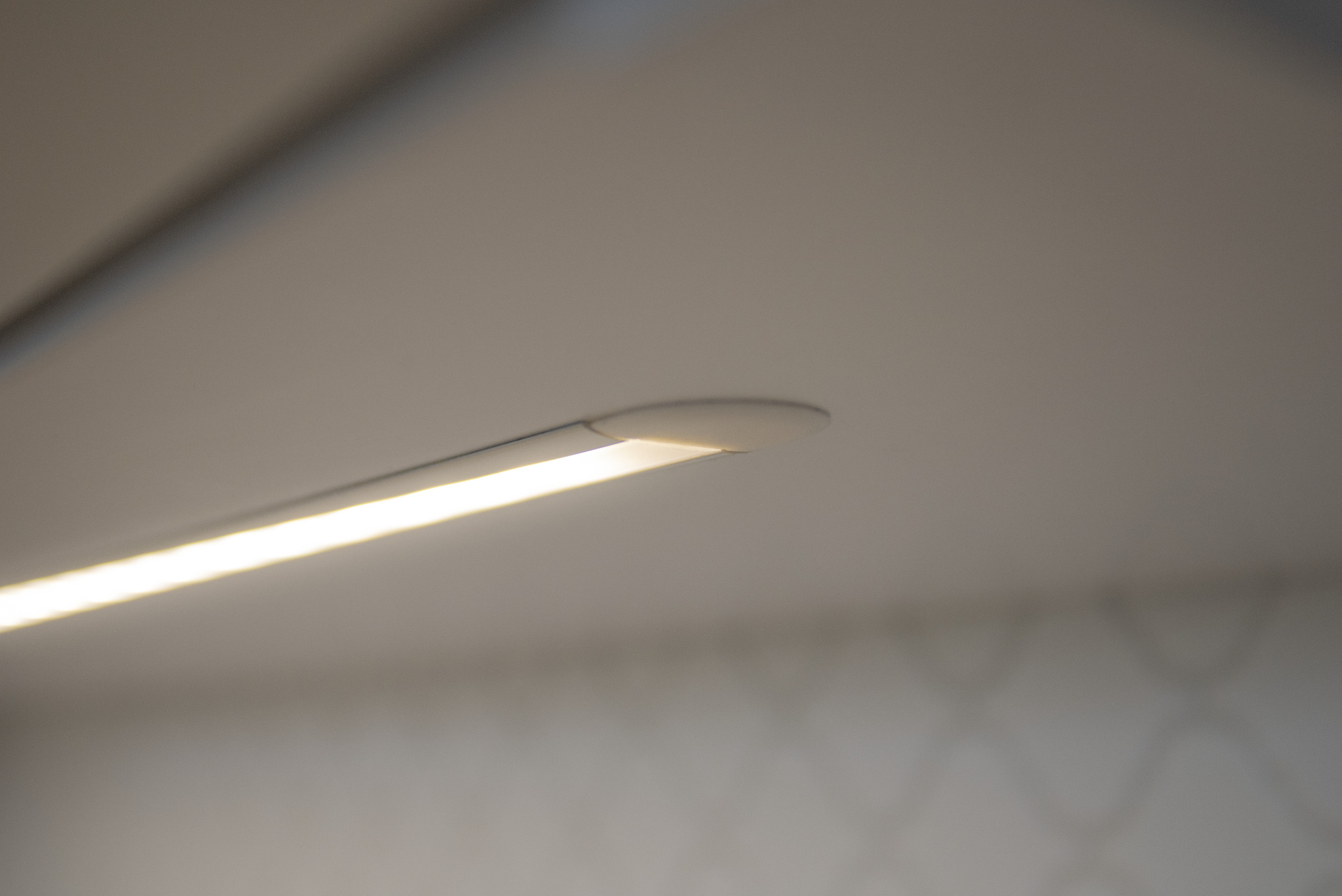 Detal minimalistycznej lampy LED podszafkowej, emitującej ciepłe, jasne światło. Widoczne delikatne wzory na ścianie w tle, podkreślające nowoczesny design.