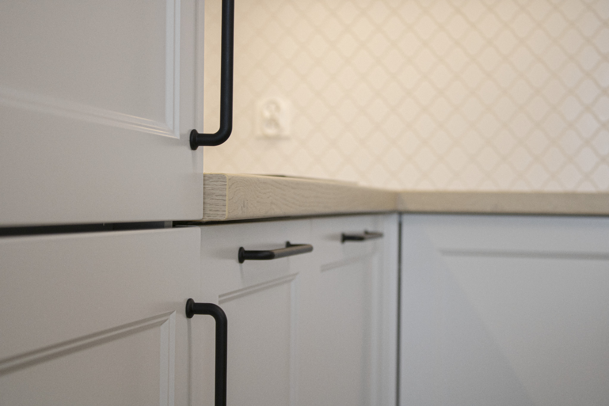 Biała zabudowa kuchenna z drewnianym blatem i czarnymi uchwytami. Minimalistyczny design z subtelnym wzorem na ścianie w tle.