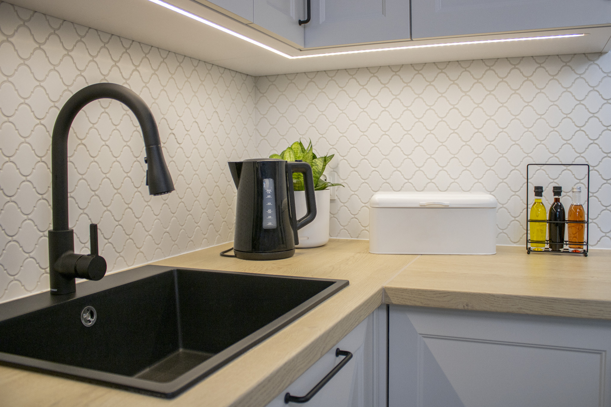 Nowoczesna kuchnia: czarny zlew, bateria, drewniany blat i białe płytki mozaikowe. Oświetlenie LED podkreśla detale.
