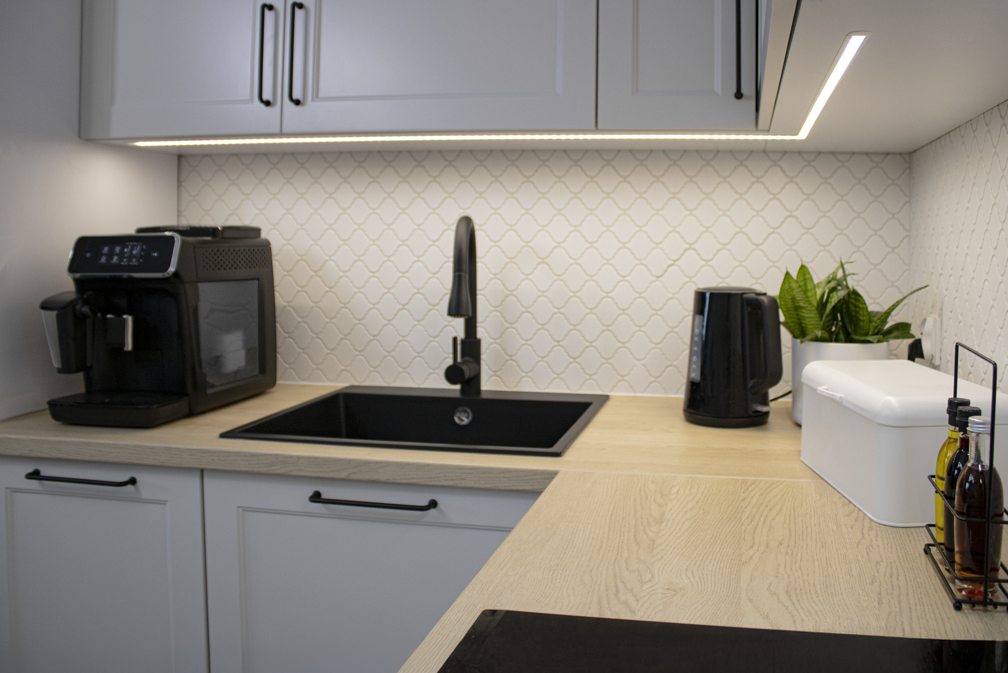 Jasna kuchnia z drewnianym blatem, czarnym zlewem i baterią. Nad blatem białe szafki z czarnymi uchwytami, podświetlenie LED i mozaika na ścianie.