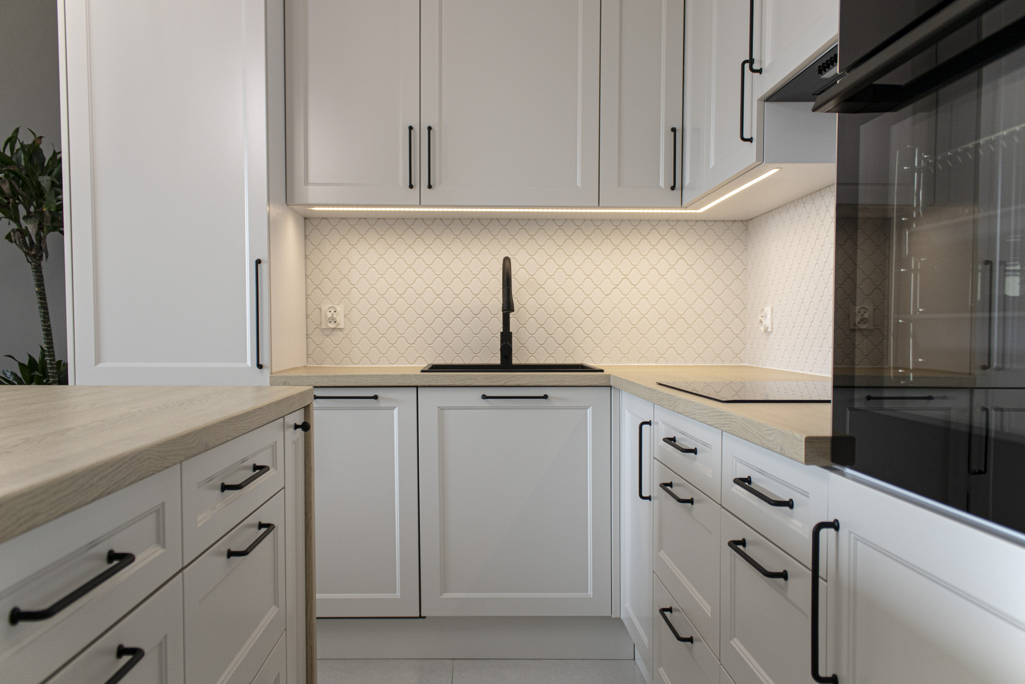 Biała kuchnia z drewnianym blatem, czarną armaturą i uchwytami. Nad blatem białe płytki w marokański wzór i oświetlenie LED. Nowoczesny design.