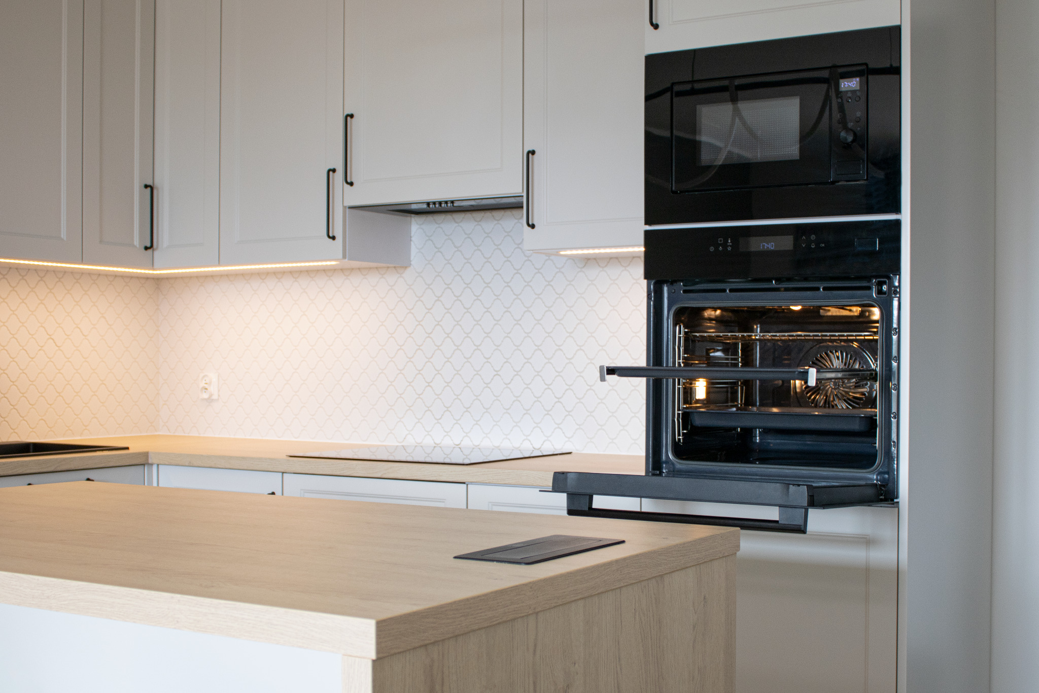 Jasna kuchnia z wyspą i zabudowaną piekarnikiem oraz mikrofalówką. Fronty szafek białe, uchwyty czarne. Na blacie płyta indukcyjna i gniazdko.