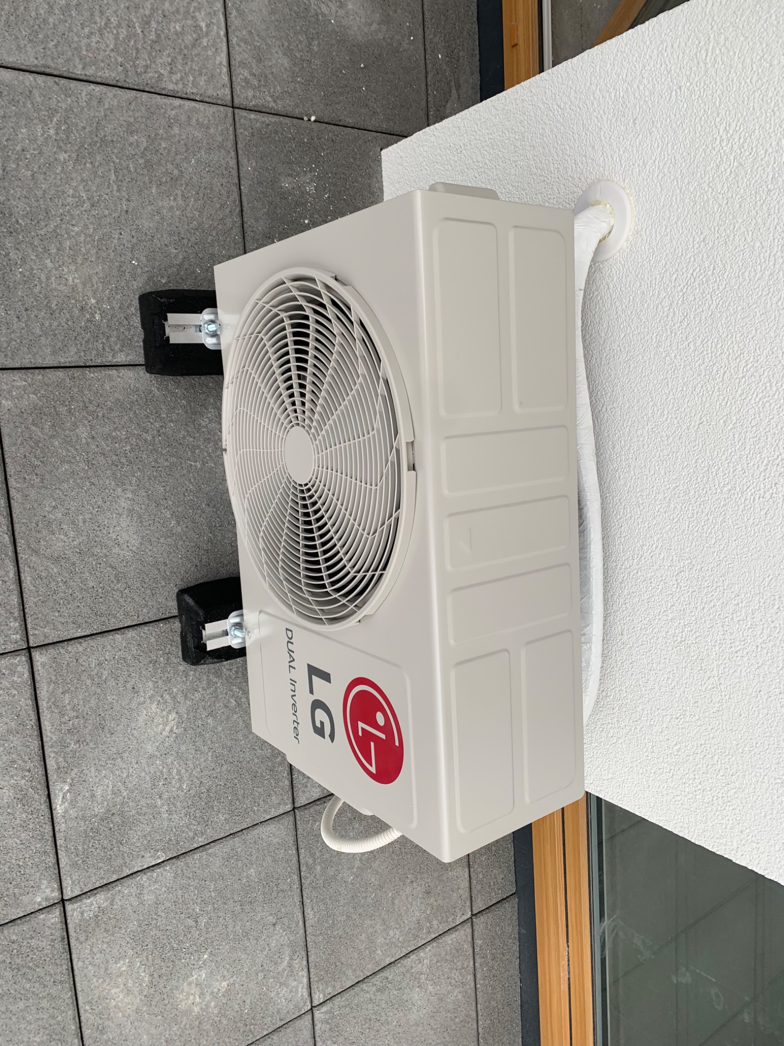 Zewnętrzna jednostka klimatyzatora LG Dual Inverter zamontowana na ścianie budynku, widoczne przewody w peszlu, czarne podkładki antywibracyjne pod wspornikami, szara kostka brukowa na podłożu.