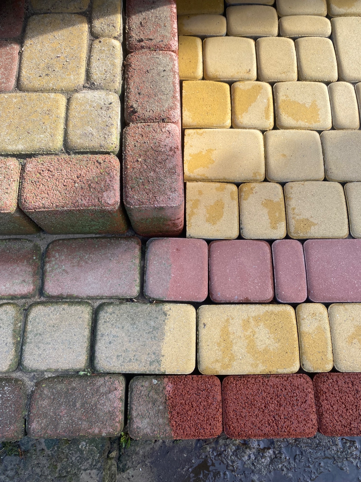 Kolorowa kostka brukowa ułożona w geometryczny wzór, częściowo mokra, z widocznymi różnicami w odcieniach i strukturze poszczególnych elementów.