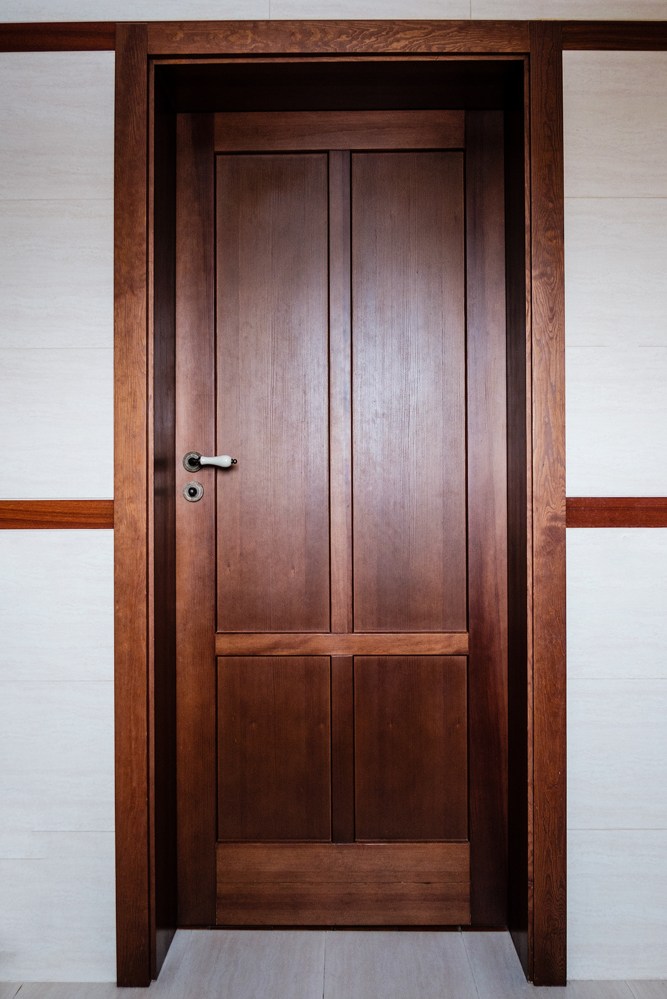 Ciemnobrązowe drzwi wewnętrzne z drewnianą ościeżnicą, widoczne w Wieliszewie, z białą klamką i zamkiem na tle białej ściany z poziomym drewnianym pasem.