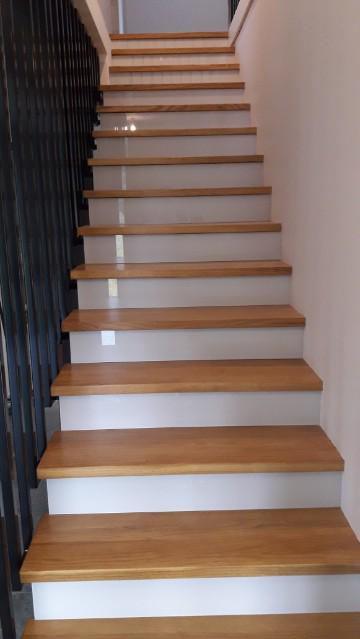 Bieg schodów z drewnianymi stopniami o ciepłym odcieniu, wspartych na białych podstopnicach i prowadzących w górę, z czarną, metalową balustradą po lewej stronie.