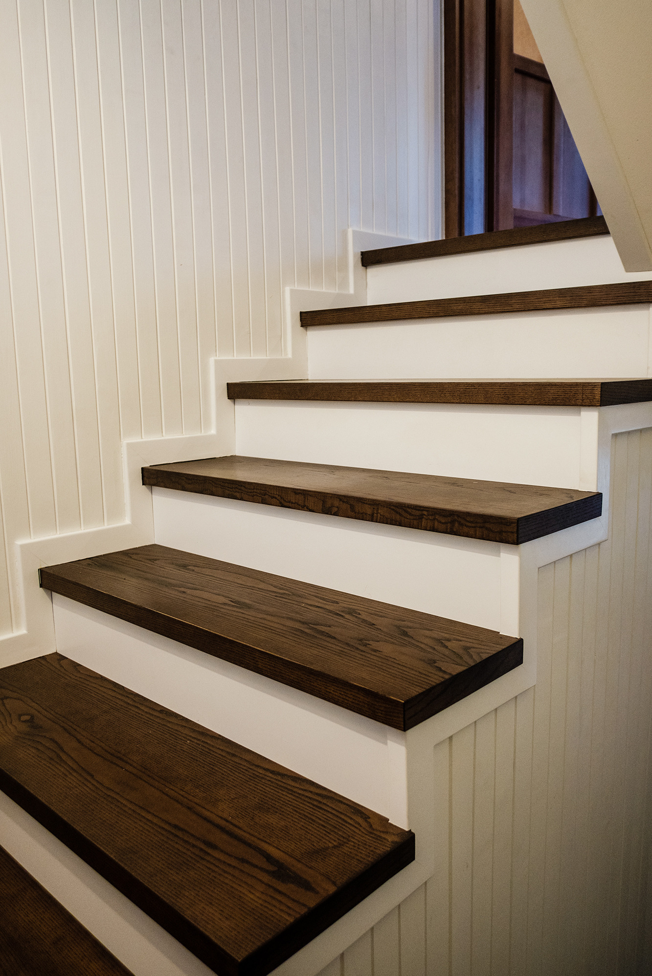 Fragment nowoczesnych schodów z ciemnego drewna, z białymi podstopnicami i panelami ściennymi w pionowe paski.