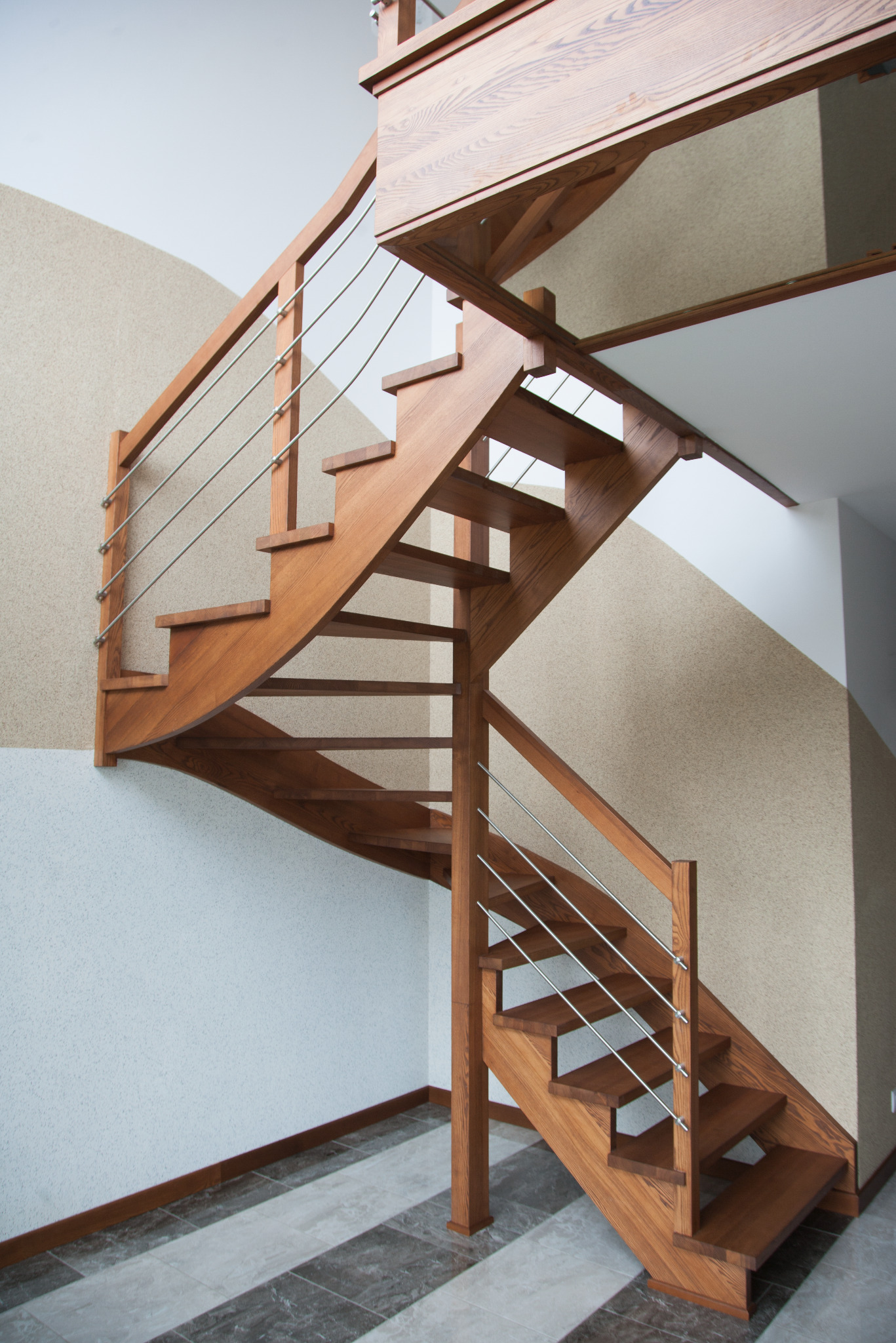 Spiralne schody z jasnego drewna z metalowymi poręczami w nowoczesnym wnętrzu z dwukolorowymi ścianami i płytkami podłogowymi.