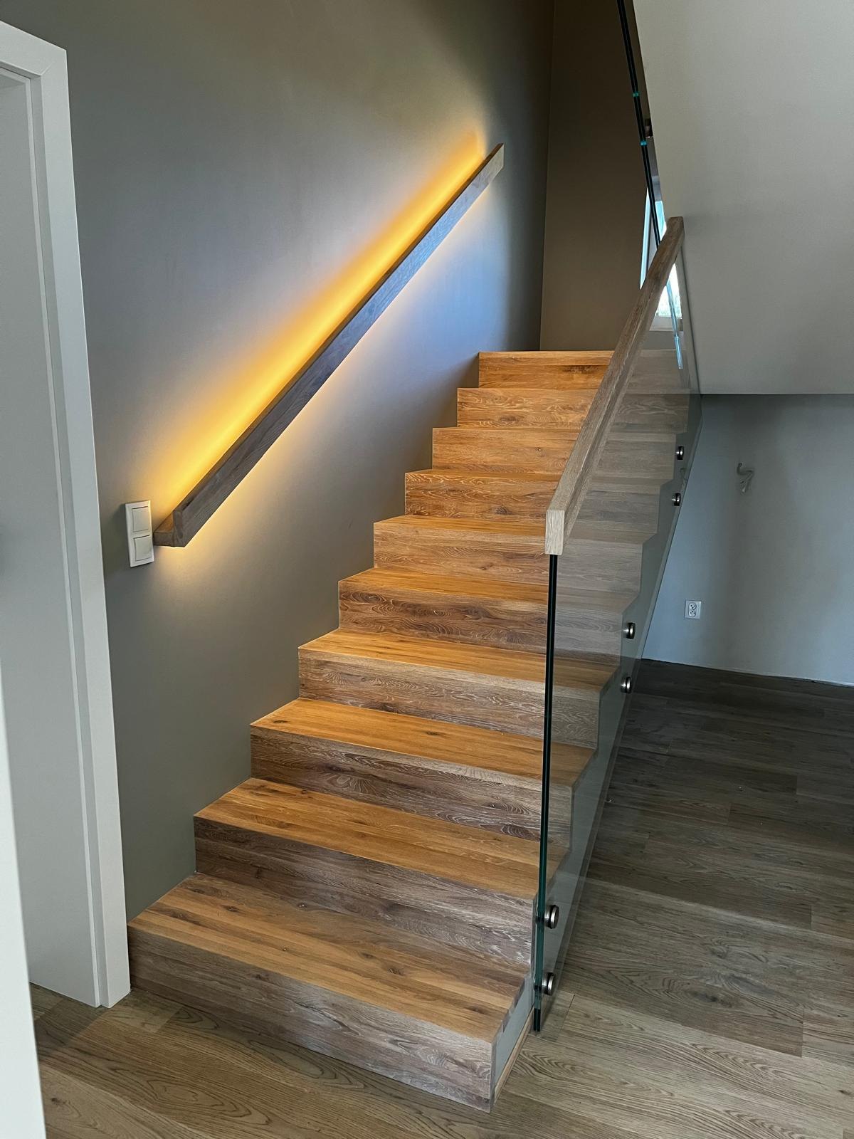 Nowoczesne schody z drewnianymi stopniami i szklaną balustradą, podświetlana poręcz na szarej ścianie, widok z dołu.