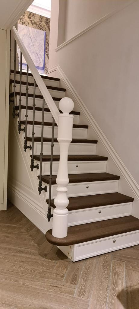 Klasyczne schody z ciemnego drewna i białymi podstopnicami, dekoracyjna balustrada z metalowymi prętami i białym słupkiem z kulą na szczycie, parkiet ułożony w jodełkę.