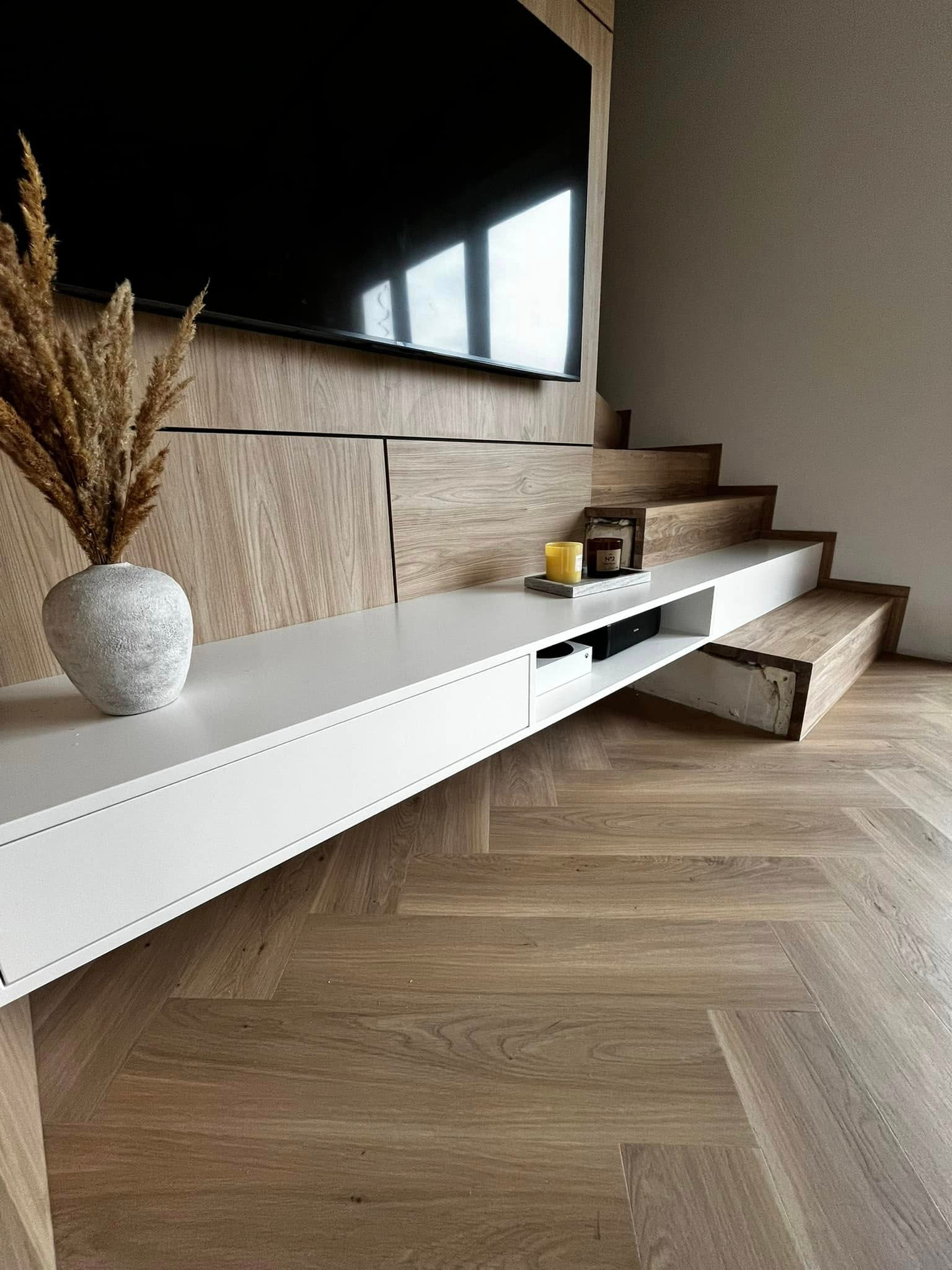 Nowoczesne schody z drewnianymi stopniami zintegrowane z zabudową telewizyjną i białą szafką RTV, parkiet w jodełkę na podłodze, dekoracyjny wazon z suszem na szafce.