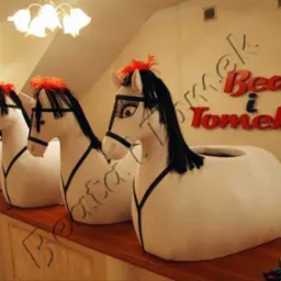 Rząd czterech ręcznie robionych, białych, pluszowych koni z czarnymi grzywami i pomarańczowymi pi&oacute;rami, ustawionych na drewnianym blacie, z napisem 'Beata i Tomek' na ścianie w tle.