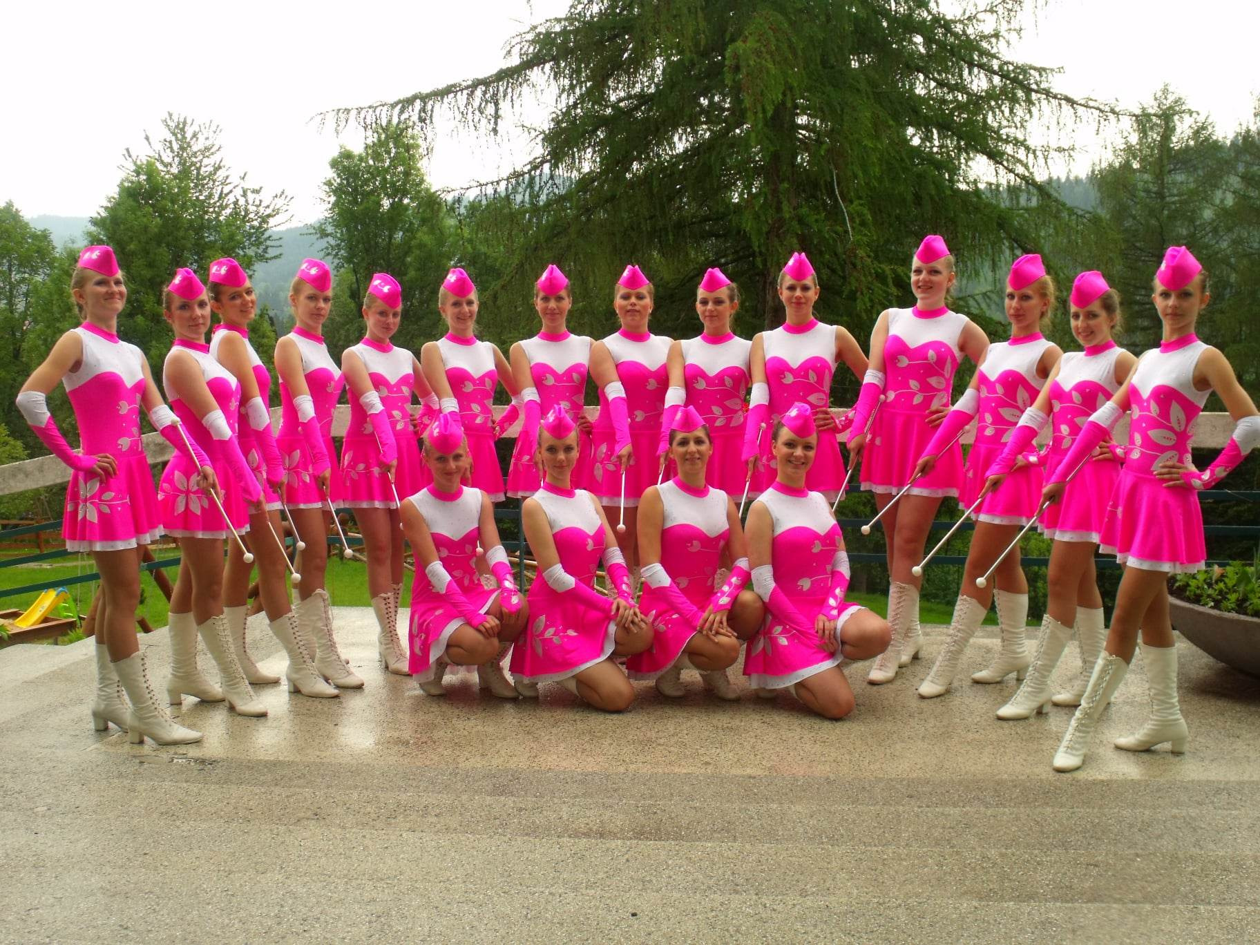 Grupa kilkunastu młodych kobiet w różowo-białych kostiumach i białych kozakach, ustawiona w dwóch rzędach, trzymających pałeczki, pozujących na zewnątrz na tle drzew