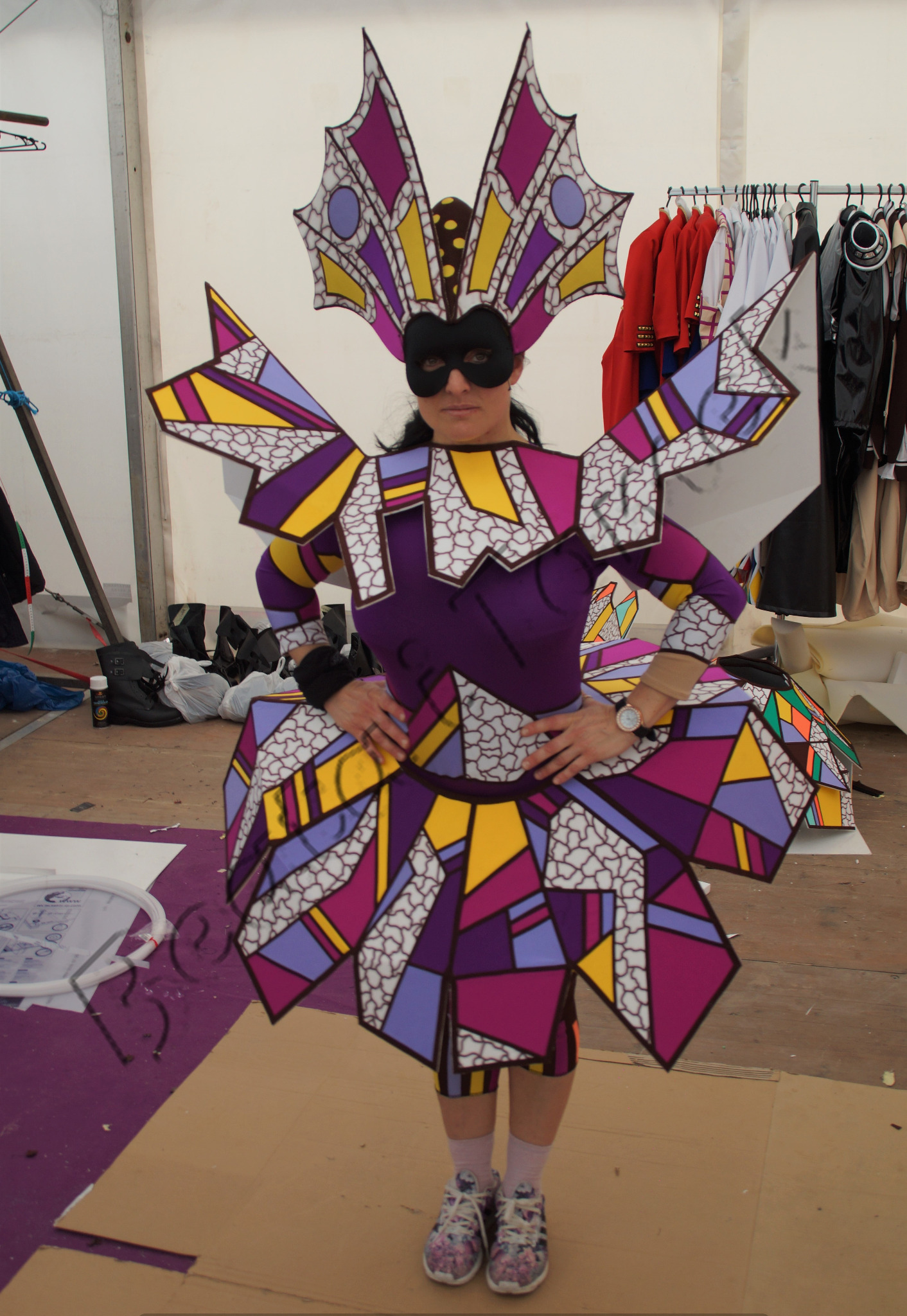 Kobieta w geometrycznym kostiumie karnawałowym z maską, stojąca w pracowni z rekwizytami i ubraniami w tle.