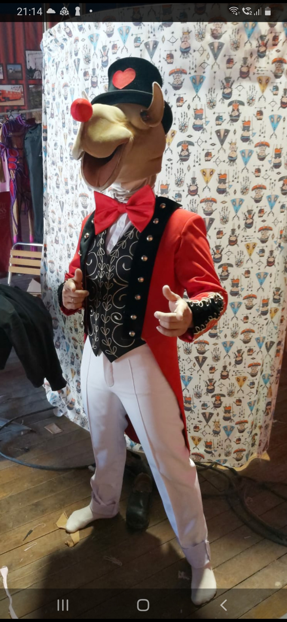 Manekin w stroju konferansjera cyrkowego z głową zwierzęcia, czerwonym nosem i cylindrem z sercem, prezentujący strój na tle wzorzystej tkaniny.