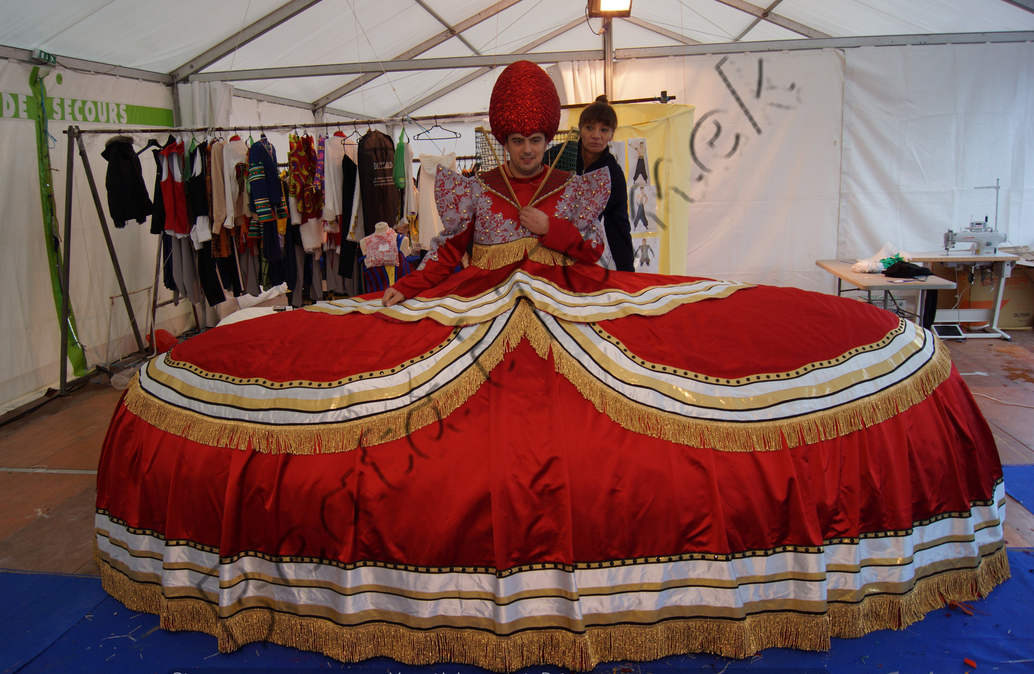 Mężczyzna w obszernym, czerwonym kostiumie balowym z dekoracyjnymi frędzlami, stojący w namiocie z wieszakami pełnymi ubrań i maszyną do szycia w tle.