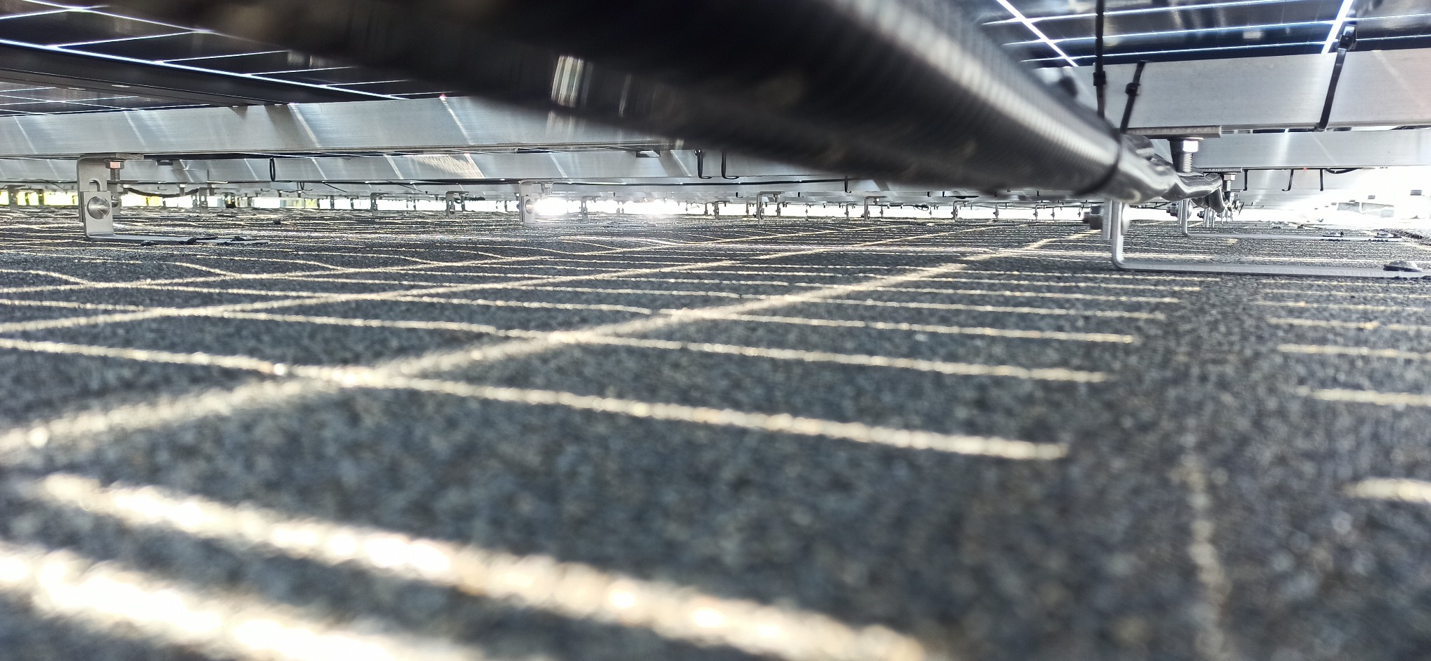 Nietypowe ujęcie od spodu paneli słonecznych, widoczna metalowa konstrukcja wsporcza z mocowaniem, perspektywa z dołu ukazuje wzór paneli w tle z silnymi refleksami słonecznymi.