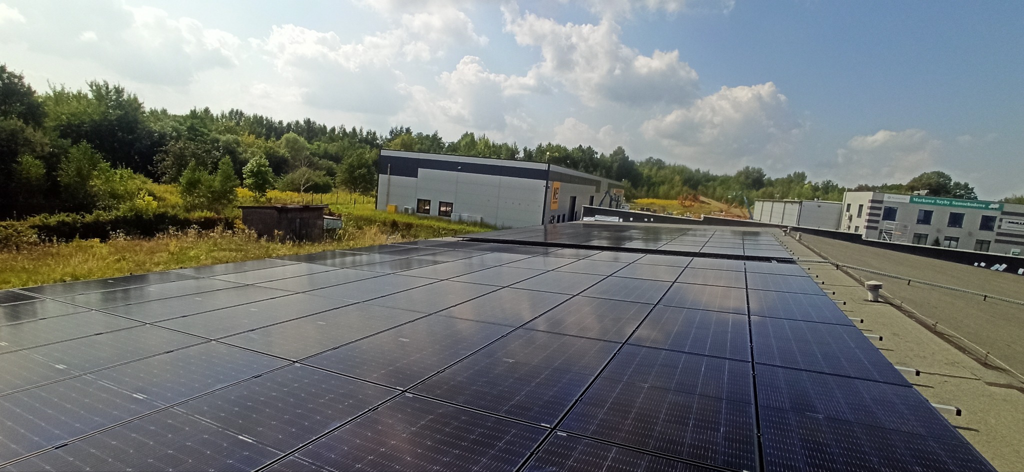 Panele fotowoltaiczne na dachu budynku przemysłowego, widok z góry z lekkim kątem, w tle zieleń drzew i budynki biurowe w słoneczny dzień.