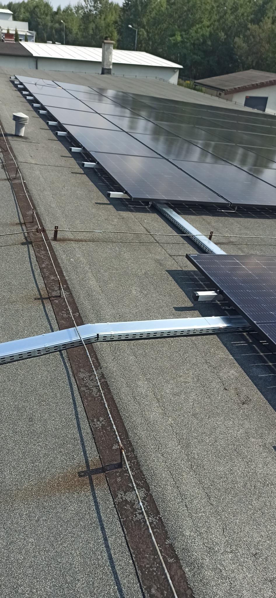Rząd paneli słonecznych na płaskim dachu budynku przemysłowego, widoczne metalowe konstrukcje wsporcze i uziemienie.