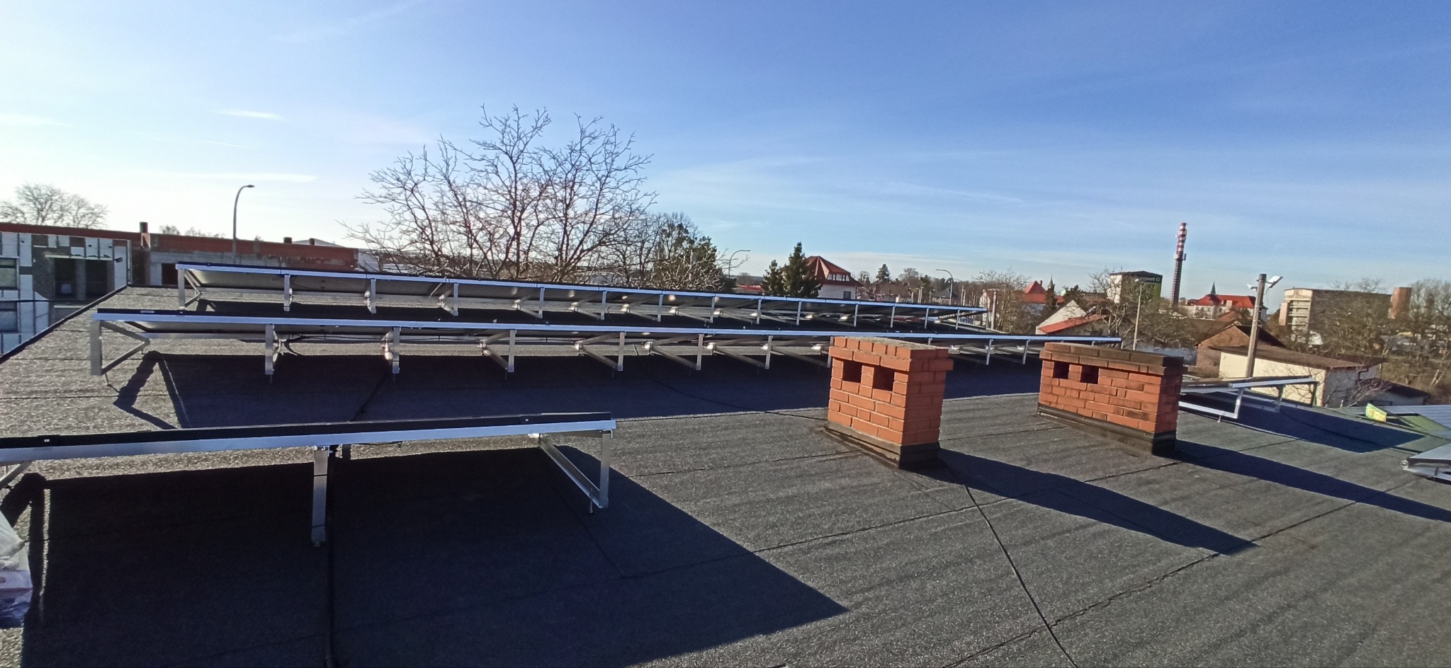 Stelaż pod panele słoneczne zamontowany na dachu pokrytym papą, widoczne dwa kominy z cegły i panorama miasta w tle, bez paneli.