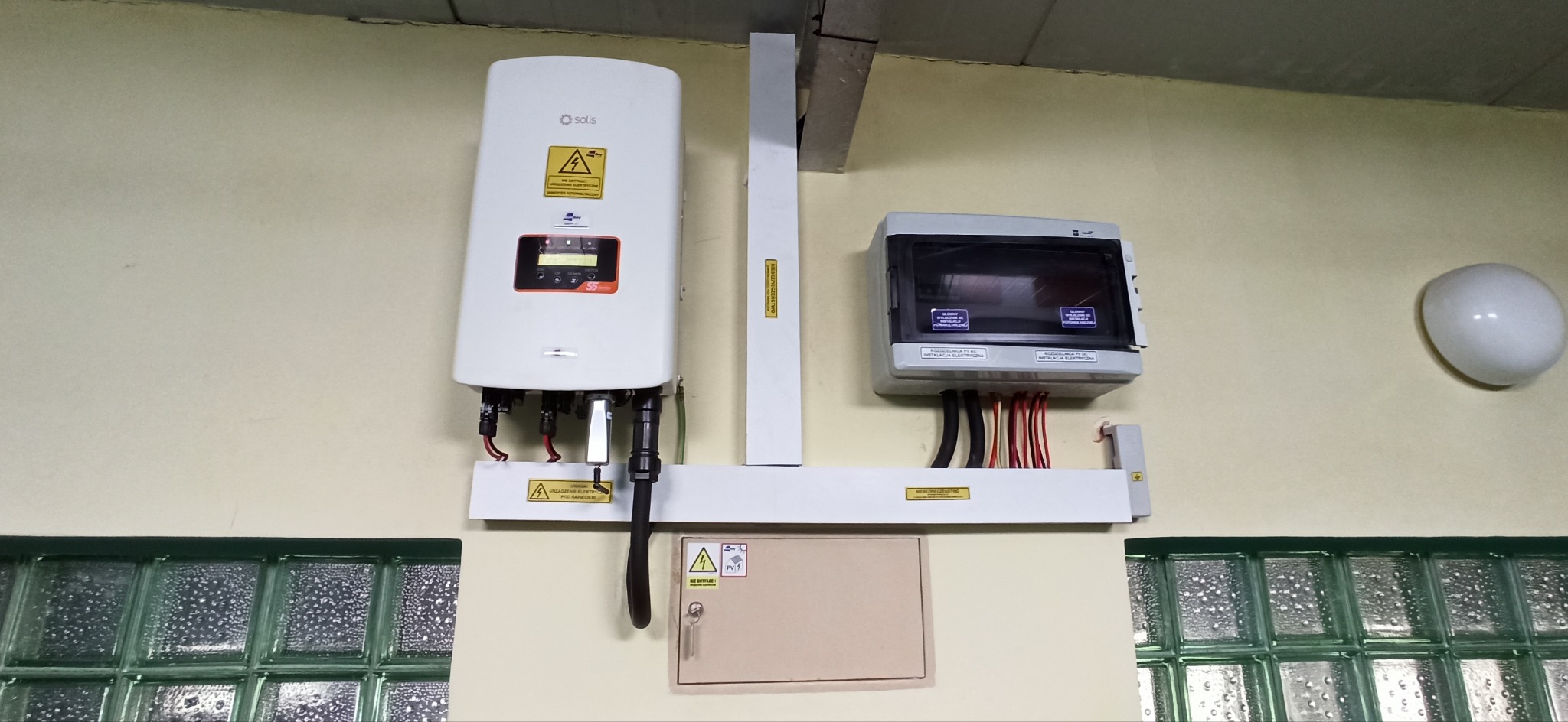 Zamontowany inwerter solarny Solis S5 z podłączonymi kablami, obok skrzynka elektryczna z zabezpieczeniami, widoczne okno z luksferami i lampa sufitowa.