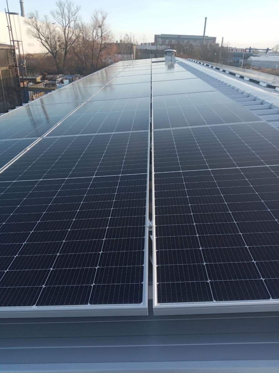 Rząd paneli słonecznych na płaskim dachu przemysłowym w słoneczny dzień, widoczne elementy montażowe i tło z zabudowaniami.