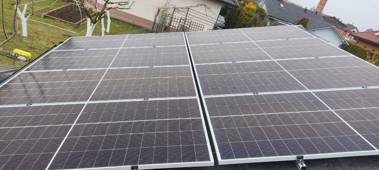 Dwa rzędy paneli słonecznych zamontowanych na dachu budynku, widok z góry na tle zabudowy jednorodzinnej i ogrodu z białymi pniami drzew.