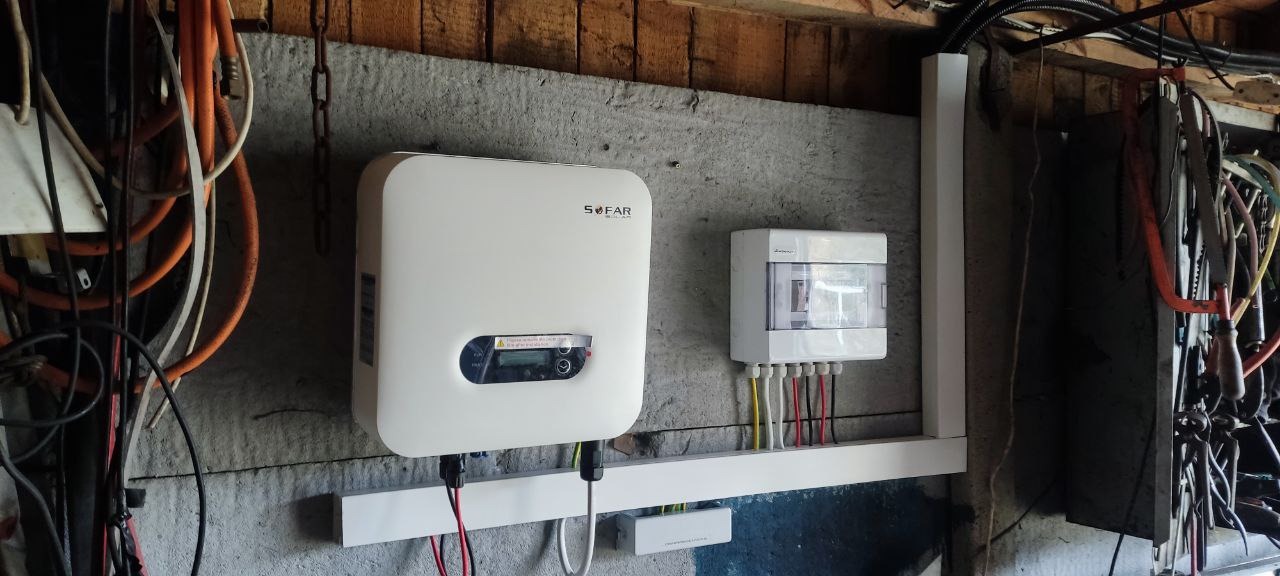 Biały inwerter Sofar Solar i skrzynka elektryczna zamontowane na szarej, betonowej ścianie wewnątrz budynku o drewnianej konstrukcji, widoczne kable i narzędzia.